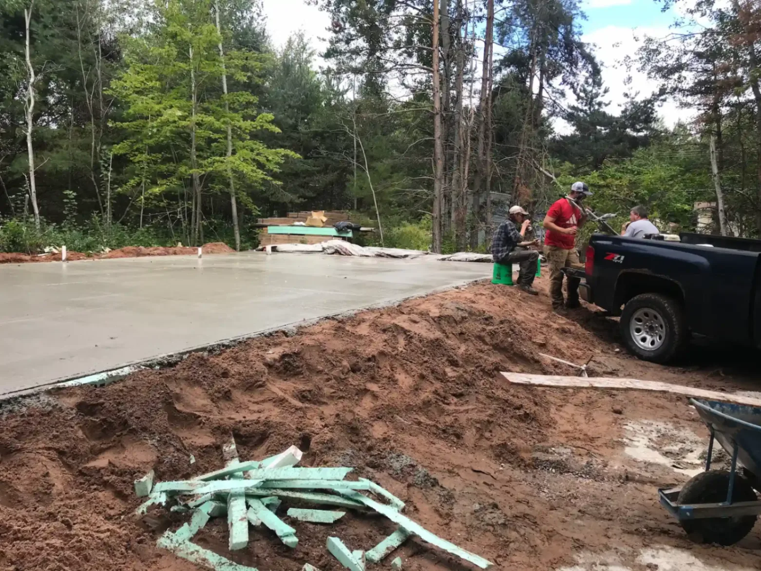 Freshly poured and finished concrete slab with crew standing beside a pickup truck on red clay soil in Marquette, Michigan